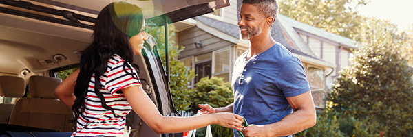 A woman with long hair stands in front of an open SUV trunk and hands full bags over to a man.